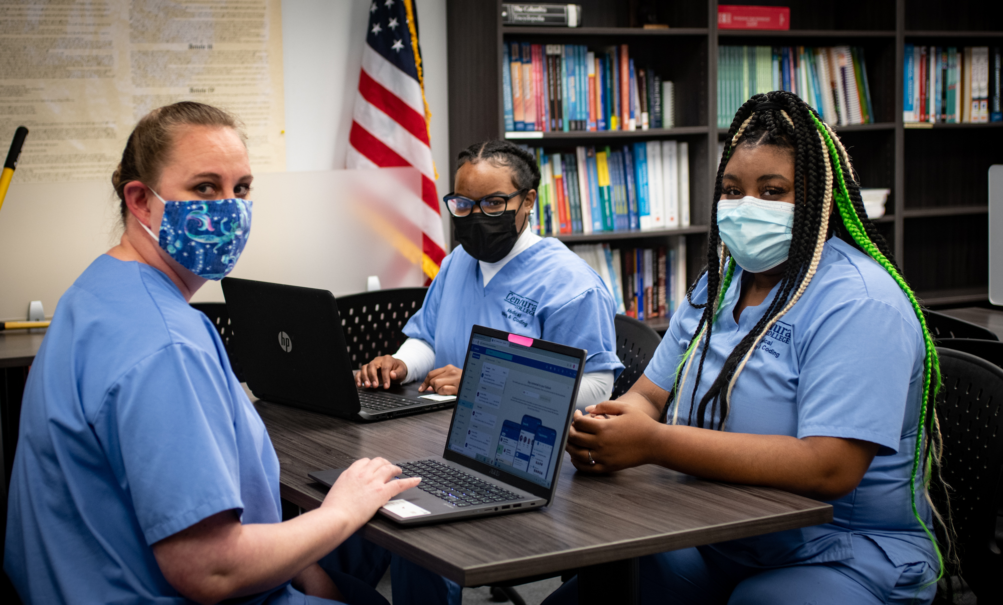 3 female medical billing and coding students at Centura College smile underneath their masks. Sitting at a table with laptops in front of them.